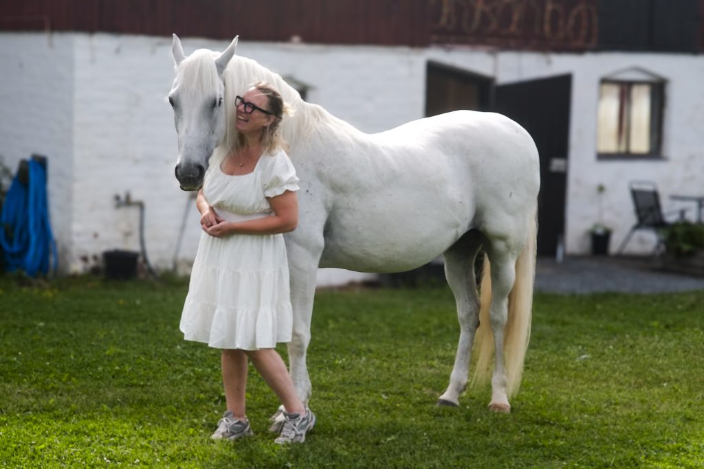 Annika Lif, iförd vit klänning, står bredvid Lipizzanerstoet Musica.
